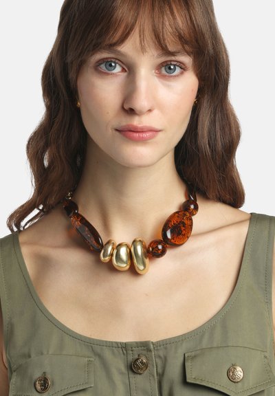 Stevige ketting met barnsteenkleurige harskralen en goudkleurige accenten in verschillende organische vormen, gedragen door een model met een groene top.