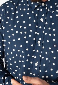 Camicia blu navy con bottoni a tinta unita e pois bianchi, dotata di colletto classico, taschino frontale e bottoni bianchi. Texture liscia.