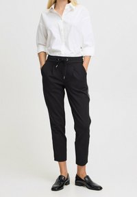 Mustat kapenevat juoksuhousut, joissa on kiristysnauhavyötärö ja sivuraitoja, yhdistettynä valkoiseen nappipaitaan ja mustiin loafereihin.