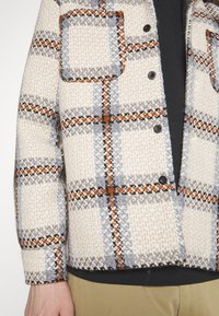 Texturerad rutig jacka i beige, grått och rostfärg; har två fickor, svarta knappar och en design med åtsittande ärmslut.