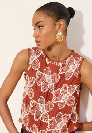 Woman with dark hair in a bun wearing a rust sleeveless top with large white floral patterns and large gold textured earrings.