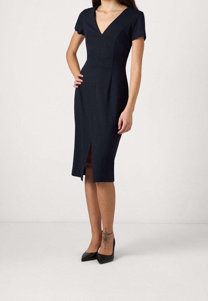 Femme portant une robe bleu marine ajustée jusqu'aux genoux avec manches courtes, col en V, fente à l'avant, talons noirs pointus et un tatouage à la cheville sur la jambe gauche.
