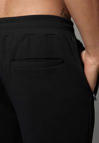 Zwarte elastische sportbroek met een elastische tailleband, voorzien van een achterzak en een zijvak met rits voor opbergruimte.