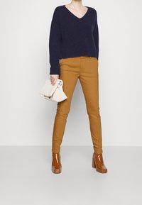 Marinblå stickad tröja med v-hals, senapsgula slim-fit byxor och glänsande bruna klackstövlar. Modell som håller i en krämfärgad handväska.