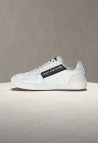 Λευκό sneaker χαμηλού προφίλ με οπές, μαύρη ρίγα και κείμενο "Versace Jeans Couture" στο πλάι, τοποθετημένο σε ουδέτερη επιφάνεια.