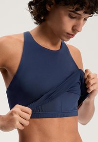 Une personne soulève l'avant d'un débardeur de sport sans manches bleu marine pour révéler une couche de soutien en maille intégrée en dessous.
