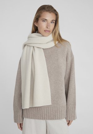 Pull côtelé beige avec un large décolleté et des manches longues, associé à une écharpe côtelée douce et claire beige drapée autour du cou.