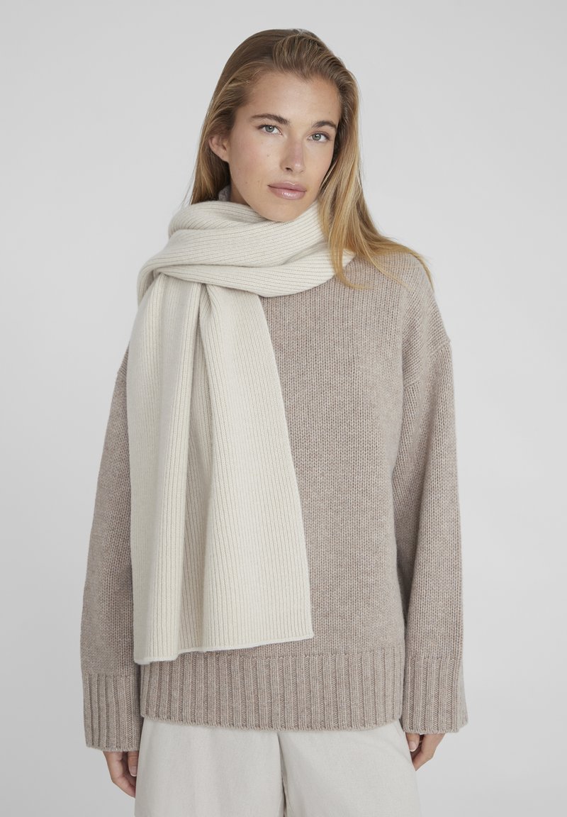 Maglione beige a coste con scollo ampio e maniche lunghe, abbinato a una morbida sciarpa beige chiaro a coste, drappeggiata intorno al collo.