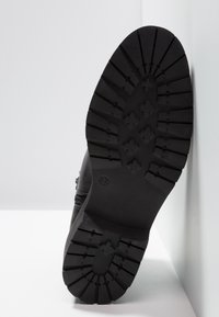 Semelle de botte noire avec motifs de profonde bande de roulement et marquage taille 37, positionnée à la verticale contre des murs blancs.