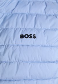 Svetlo modra napihnjena jakna ima šivane panele, gladko teksturo in izstopajoč črn logotip "BOSS" na levi strani.