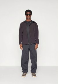 Zwarte jas met ritssluiting over een nauwsluitend zwart topje, gecombineerd met losse donkere broek en grijze sneakers, allemaal met een strak, modern ontwerp.