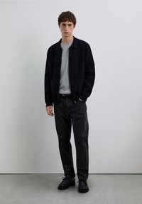 Marineblaue Wolljacke über einem grauen Hemd, kombiniert mit schwarzen Jeans und schwarzen Schnürschuhen. Das Modell steht vor einem neutralen Hintergrund.