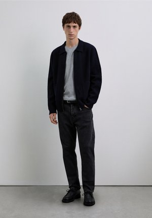 Giacca in lana blu navy sopra una camicia grigia, abbinata a jeans neri e scarpe nere stringate. Il modello è in piedi contro uno sfondo neutro.