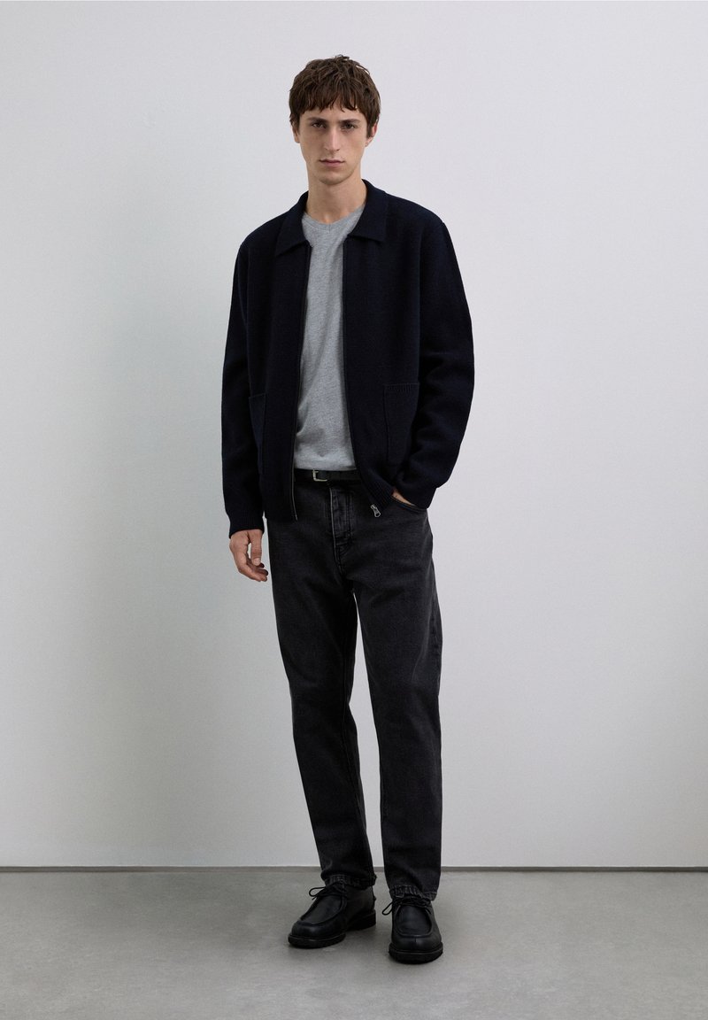 Marineblaue Wolljacke über einem grauen Hemd, kombiniert mit schwarzen Jeans und schwarzen Schnürschuhen. Das Modell steht vor einem neutralen Hintergrund.