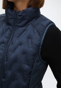 Μπλε επενδυμένο γιλέκο με ψηλό γιακά, που διαθέτει φερμουάρ, αντίθετη ραφή και υφασμάτινες πλευρές με υφή. Λείο ύφασμα με ματ φινίρισμα.