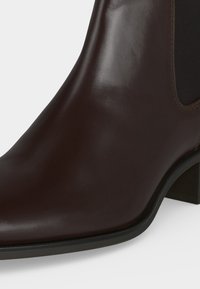 Κοντινή λήψη ενός σκούρου καφέ δερμάτινου μποτάκι με στρογγυλεμένη μύτη, μαύρη ελαστική πλαϊνή επένδυση και χαμηλό χοντρό τακούνι σε λευκό φόντο.