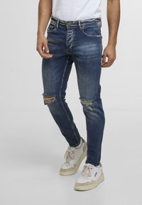 Jeans din denim albastru, fit skinny, cu gauri distorsionate la genunchi, accente estompate și închidere cu buton. Asortați cu adidași albi și bej.