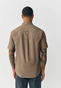 Korte mouwen bruine shirt met een rugplooitje, knoopsluiting en een gladde textuur; gedragen met donkere broeken en zichtbare getatoeëerde armen.