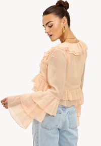 Femme aux cheveux bruns attachés en chignon, portant des boucles d'oreilles créoles dorées, un chemisier transparent pêche à volants avec des manches évasées, et un jean en denim bleu clair.