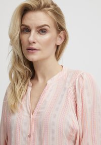 Blusa rosa chiaro con scollo a bottone, caratterizzata da strisce testurizzate in tonalità di rosa scuro e motivi bianchi. Tessuto morbido e fluido.
