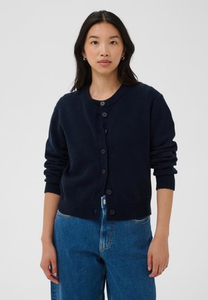 Femme aux longs cheveux foncés portant un cardigan bleu marine boutonné et un jean bleu, debout devant un fond gris clair uni.