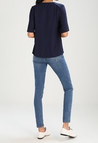 Blouse bleu marine à manches courtes, détail en partie haute du dos, associée à un jean skinny bleu clair et des chaussures blanches à lacets. Texture de tissu lisse.