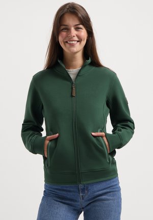 Smiling woman wearing a dark green zip-up jacket with hands in pockets and blue jeans, standing against a plain light background.