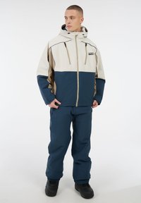 Winterjas en broek in beige, marineblauw en tan, gemaakt van waterafstotend materiaal. Heeft een capuchon, een rits aan de voorkant en zijzakken.