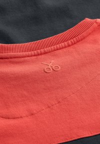 Collo del sudore corallo con texture a coste e logo a forbice, sovrapposto a un tessuto nero. Dettagli delle cuciture visibili sul collo.