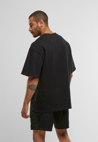 Mann mit kurzen Haaren und Tattoo am Unterarm, der ein schwarzes, übergroßes T-Shirt und schwarze Shorts trägt und seitlich vor einem schlichten Hintergrund steht.