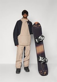 Snowboardzista ubrany w beżową i granatową wodoodporną kurtkę oraz spodnie, noszący gogle, trzymający snowboard z wiązaniami, czarne buty.