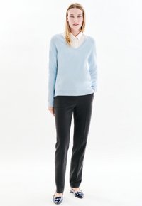 Donna in piedi con le mani nelle tasche, indossa un maglione leggero blu con scollatura a V, una camicia bianca con colletto, pantaloni scuri e ballerine nere lucide.