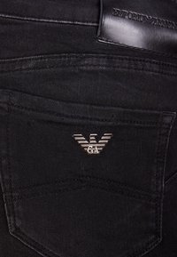 Svarta jeans av denim med en liten örnlogotyp i silver på bakfickan, och en svart läderbaksidetikett med präglad text.