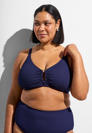 Kvinde med kort sort hår iført en marineblå tekstureret bikini, justerer stroppen, står op ad en hvid væg med skygge.