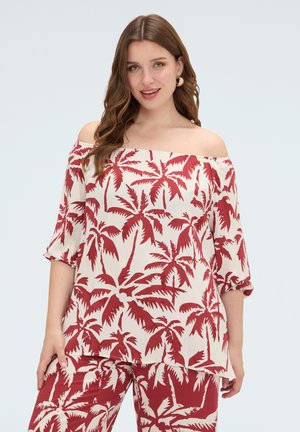 Kvinde iført off-shoulder top og matchende bukser med røde palmeprint på hvid baggrund, står og smiler.