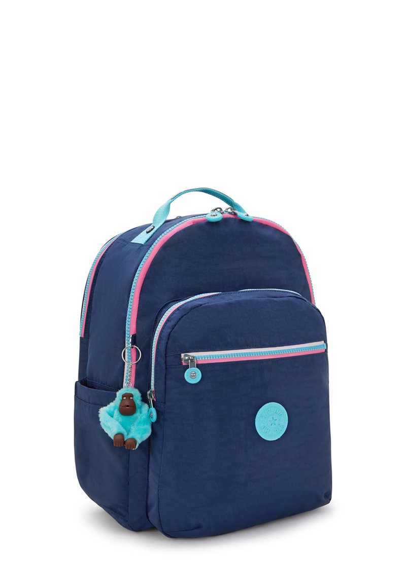Mochila de color azul marino con cremalleras rosas y turquesas, bolsillo frontal, llavero de mono de peluche y una etiqueta circular de la marca en el frente.