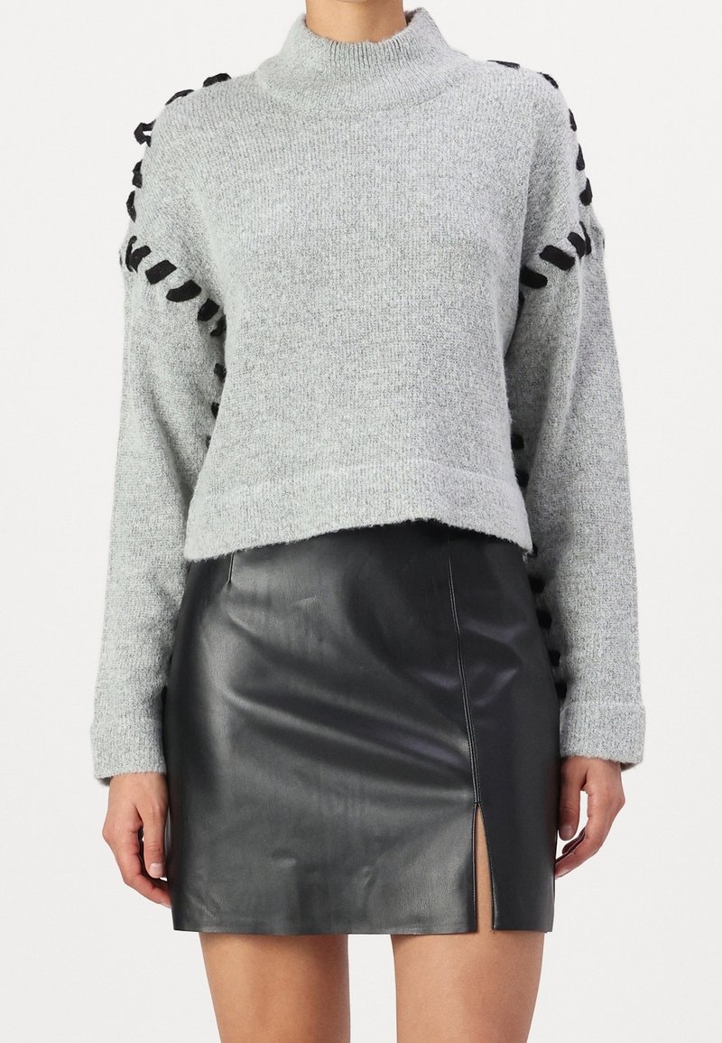 Pull gris en maille avec un col montant et des détails à lacets noirs sur les côtés, associé à une mini-jupe en cuir noire avec une fente latérale.