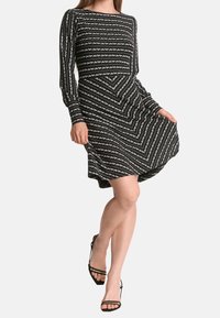 Schwarzes Kleid mit langen Ärmeln, das weiße Zickzack- und Streifenmuster aufweist. Fließendes Design mit einem taillierten Oberteil, knielang.