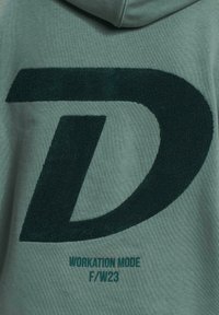 Hoodie vert arborant un grand logo "D" texturé en vert foncé et le texte "MODE DE TRAVAIL F/W23" en turquoise. Tissu doux avec une coupe décontractée.