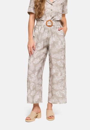 Mujer que lleva un mono beige con patrón de hojas blancas, mangas cortas, piernas anchas y un cinturón con hebilla redonda de madera, combinado con sandalias beige de tacón.