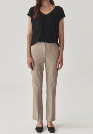 Kvinde iført sort kortærmet V-hals top, beige skræddersyede bukser med nålestriber og sorte spidse flade sko, står mod en ensfarvet baggrund.