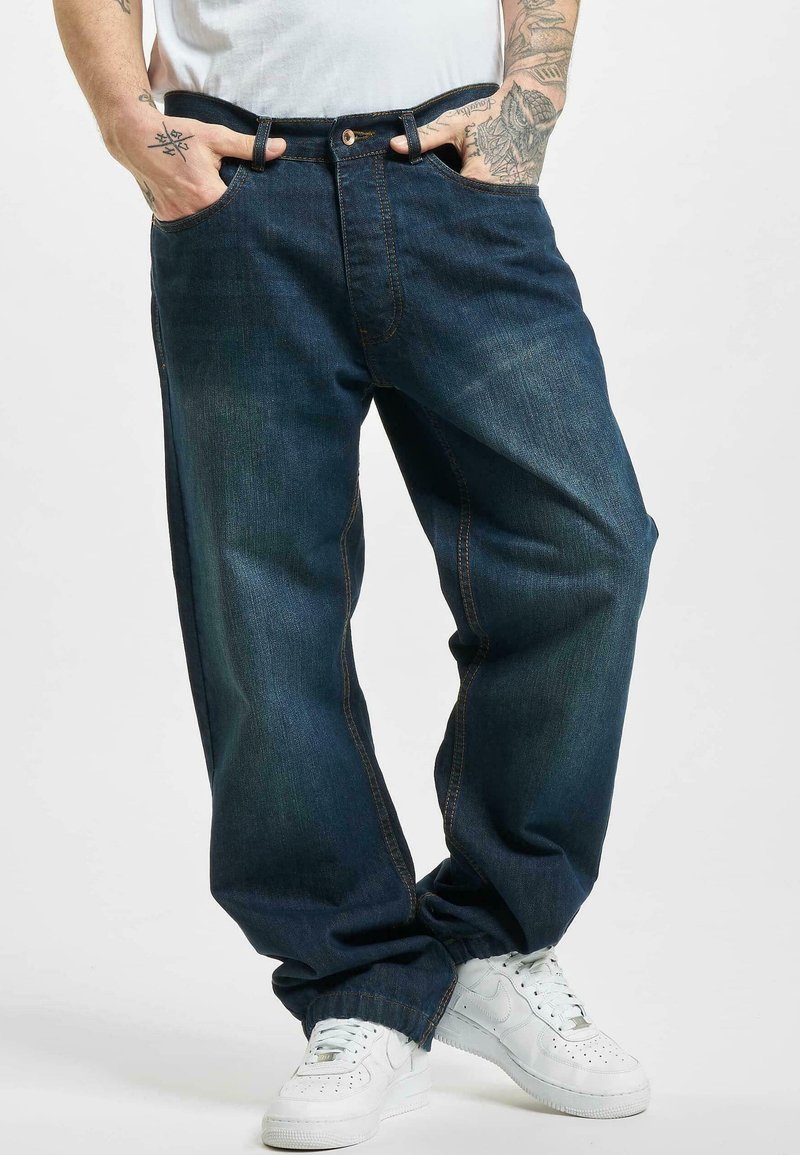 Jeans oscuros de corte holgado, confeccionados en denim, con bolsillos laterales, cremallera y costuras en un color contrastante, combinados con zapatillas blancas.