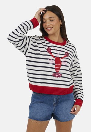 Donna che indossa un maglione a righe bianche e blu navy con rifiniture rosse e un disegno di un'aragosta rossa, abbinato a pantaloncini di jeans blu, sorridente con gli occhi chiusi.