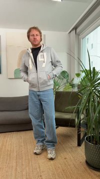 Sivá mikina na zips s bielymi detailmi, čierne tričko, svetlomodré džínsy a sivé tenisky, stojaci na tkanom koberci vedľa pohovky a rastlín.