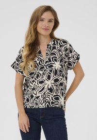 Blouse noire et crème à manches courtes, avec un motif floral, un col en V et une coupe ample, associée à un jean en denim bleu foncé.