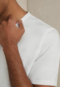 Tricou alb cu guler rotund, realizat din bumbac, cu textură netedă, design ajustat. Se văd atingeri manuale în zona gulerului, evidențiind stilul casual.
