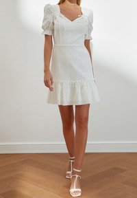 Vestido blanco con mangas abullonadas, textura de puntos, corsé ajustado y dobladillo con volantes. Combinado con sandalias blancas con tiras sobre un suelo de madera.