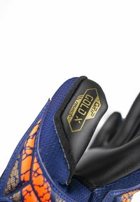 Met handschoen beklede hand met blauw en oranje ontwerp die een zwarte duimbeschermer vasthoudt met het label "Grip Gold X Gloves".