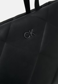Juodas odinis laikiklis su siuvinėta danga, su išdebokuotu "CK" logotipu ir tvirtais rankenomis, pabrėžiantis sklandžią tekstūrą.
