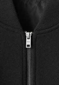 Chaqueta bomber negra con cuello acanalado, cierre frontal con cremallera y herrajes metálicos, y tejido texturizado.